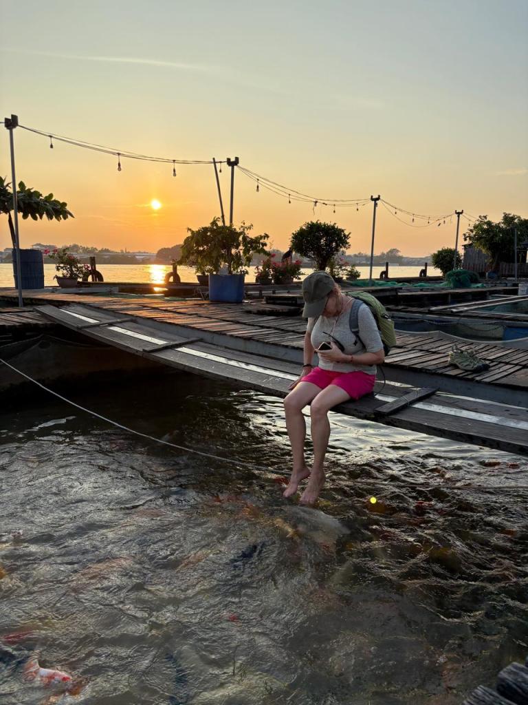 Traveler enjoying a fish massage at a floating fish farm on Son Islet, Can Tho, Vietnam, dipping feet into the Hau River at sunset.